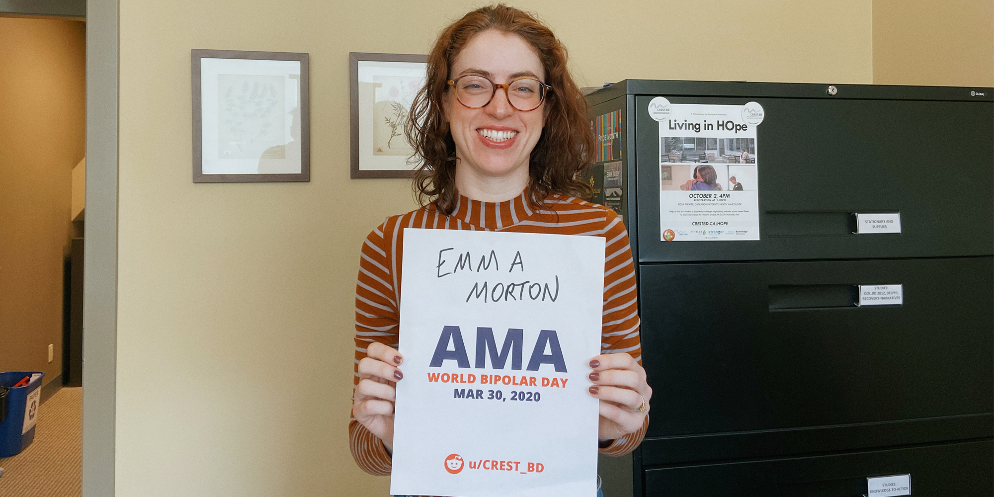 Emma in her office, smiling and holding a sign proving she will be involved in the bipolar ama.