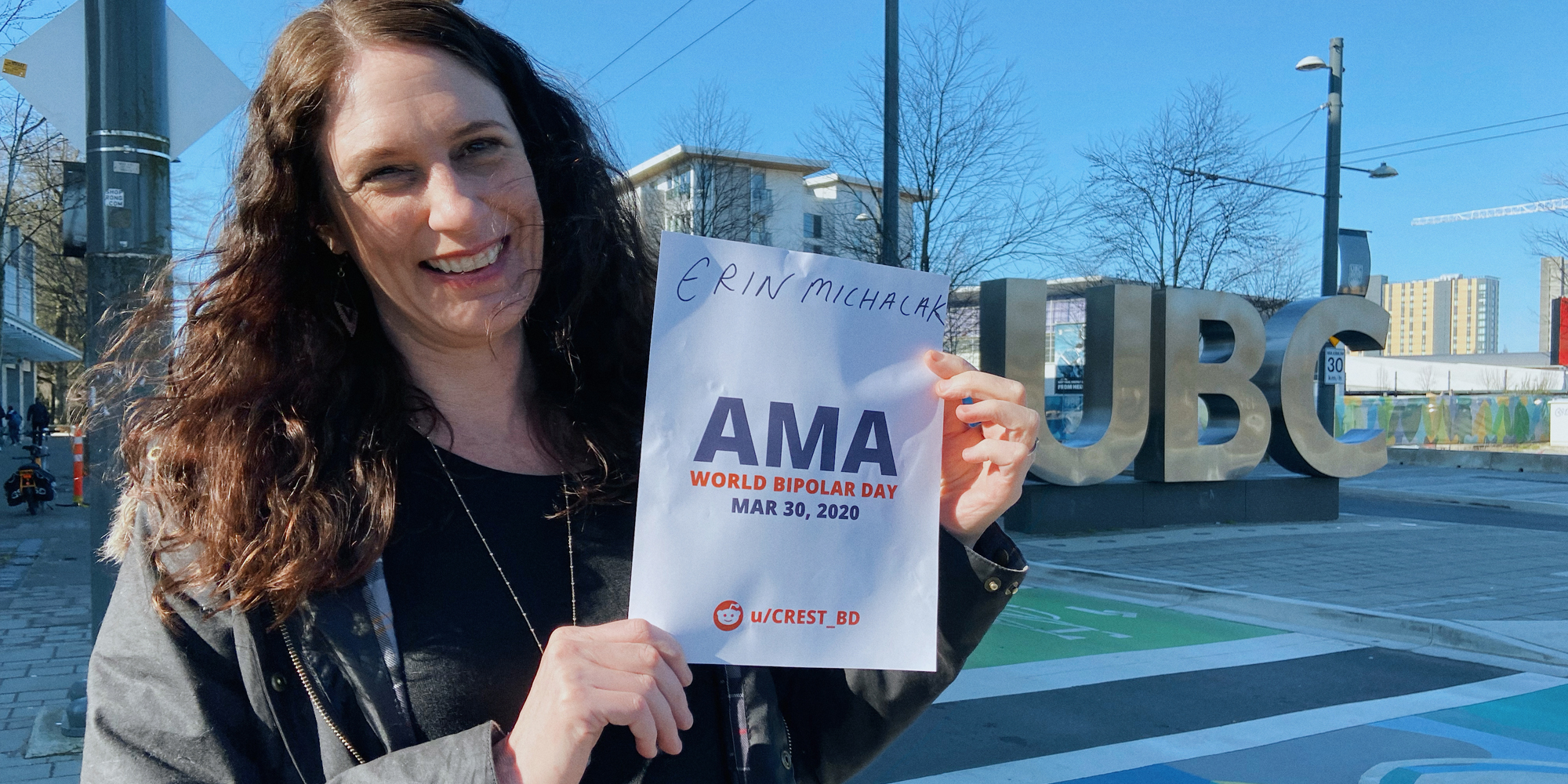 Erin outside, smiling and standing by a statue of the UBC logo. She is holding a sign proving she will be involved in the bipolar ama, with her name and the AMA date and time on it.