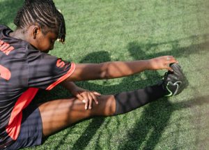 A black teenage girl wearing soccer clothes is doing a hamstring stretch on the soccer field.