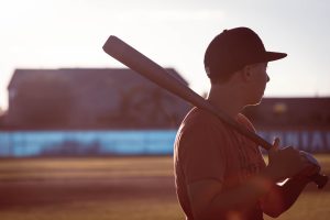 A teenage boy wearing a batting helmet and baseball jersey stands up to bat. It is sunset and he is looking away from the camera.