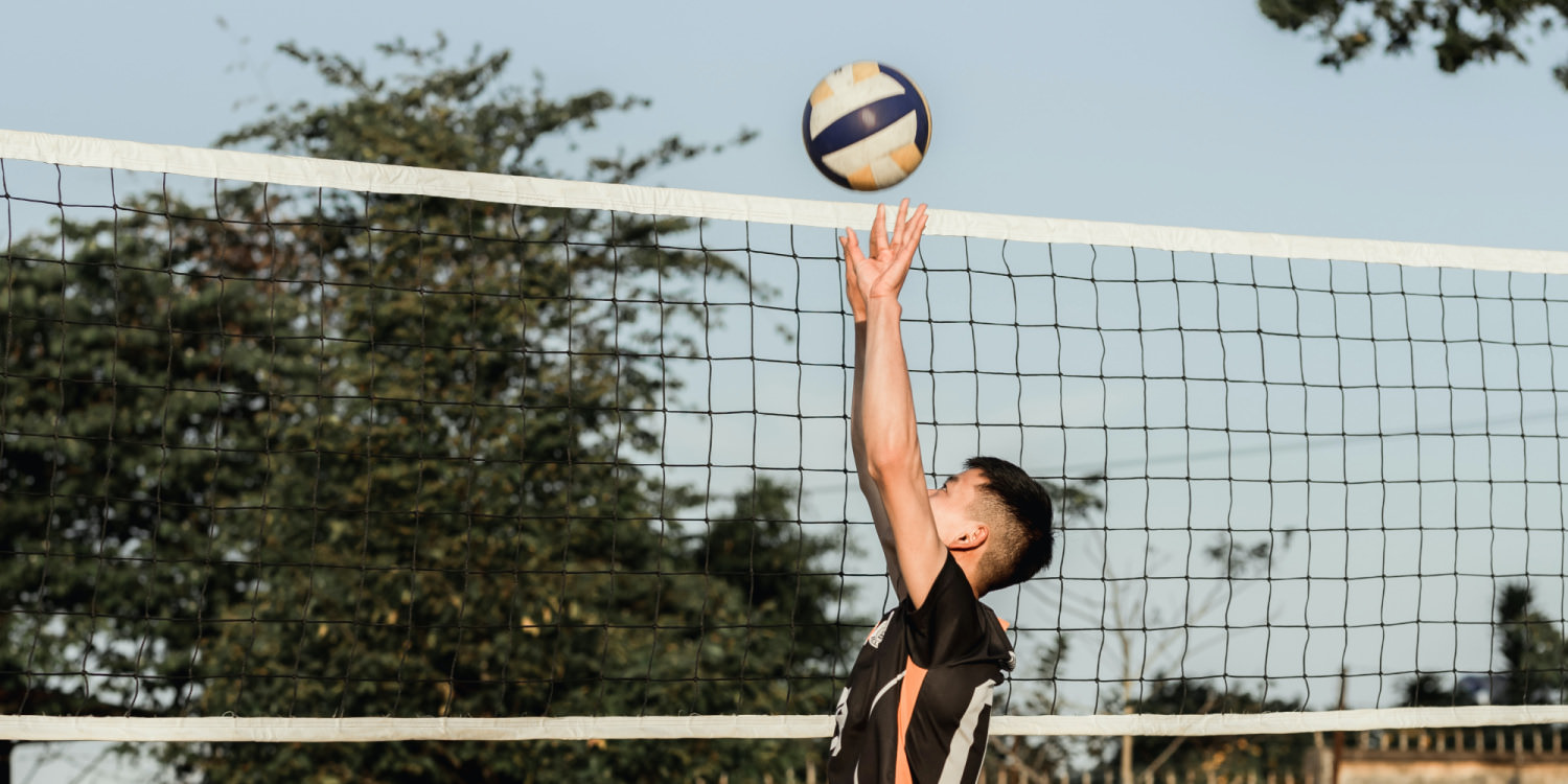 A teenage East Asian boy is playing volleyball at an outdoor court. The sky is blue and clear and there are trees nearby. He is shown in profile reaching upwards towards the ball.